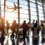 People queuing inside airport terminal with airplane flying outside.