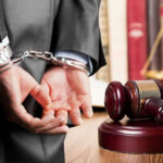 Person in handcuffs near gavel and scales of justice.