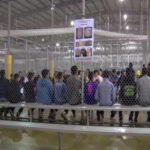 A group of young individuals seated in a detention center surrounded by security fencing and informational posters