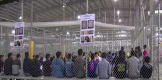 A group of young individuals seated in a detention center surrounded by security fencing and informational posters
