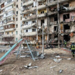 Damaged apartment building with debris scattered firefighters present.