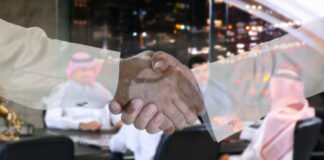 Two people shaking hands; men in background discussing.