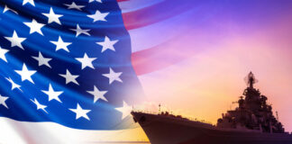 An American flag in the foreground with a naval ship silhouetted against a sunset