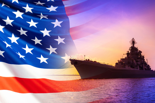 An American flag in the foreground with a naval ship silhouetted against a sunset