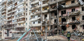 Damaged apartment building with debris scattered firefighters present.
