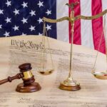 Trump Begs SCOTUS To Reconsider Birthright Decision Scales of justice and gavel on the U.S. Constitution with American flag backdrop