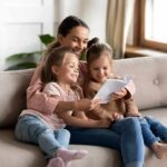 Newsom Declares 2 Muslim Holidays OFFICIAL Woman reading a book with two young girls on a sofa