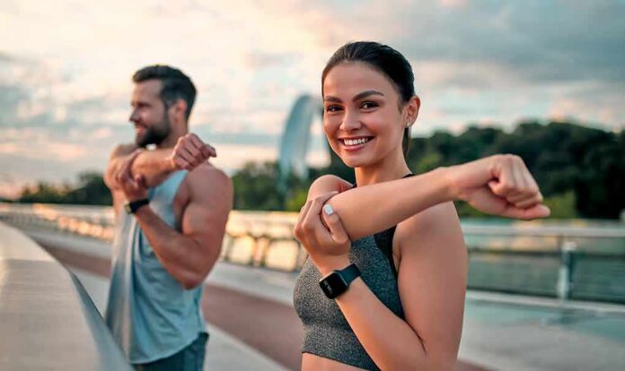 shutterstock_1779681110 Woman stretching her arm outdoors with man in background