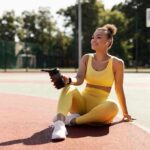Woman in yellow activewear sitting on outdoor court with water bottle