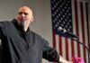 Bald man speaking at a microphone with a U.S. flag in the background