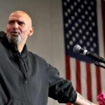 Fetterman DECLARES He’s Republican Bald man speaking at a microphone with a U.S. flag in the background