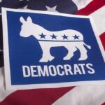 Dem Candidate CANCELED After Repeated Homophobe Slurs Democrats donkey symbol on a blue sign over an American flag