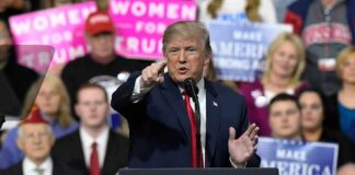Donald Trump speaking at a rally podium, pointing toward the audience