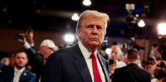 A serious-looking man in a suit with a red tie surrounded by a crowd and cameras