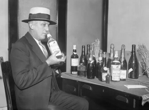 Man in a hat holding a bottle of liquor with various bottles on a table