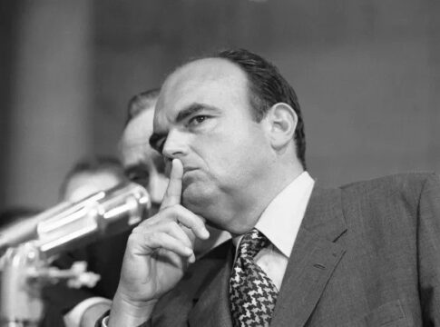 A serious man with a finger to his lips, indicating silence, during a congressional hearing