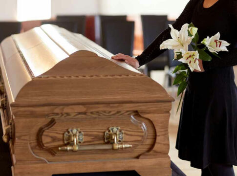 Person with lilies standing beside a wooden casket.