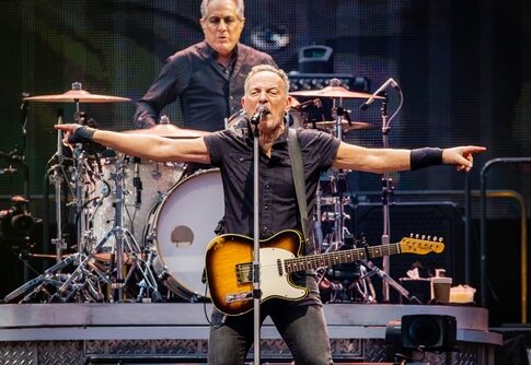 A guitarist performing passionately on stage with a drummer in the background