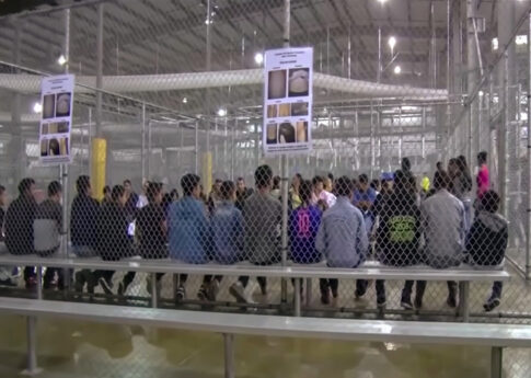 A group of young individuals seated in a detention center surrounded by security fencing and informational posters