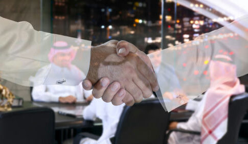 Two people shaking hands; men in background discussing.