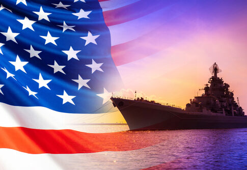 An American flag in the foreground with a naval ship silhouetted against a sunset